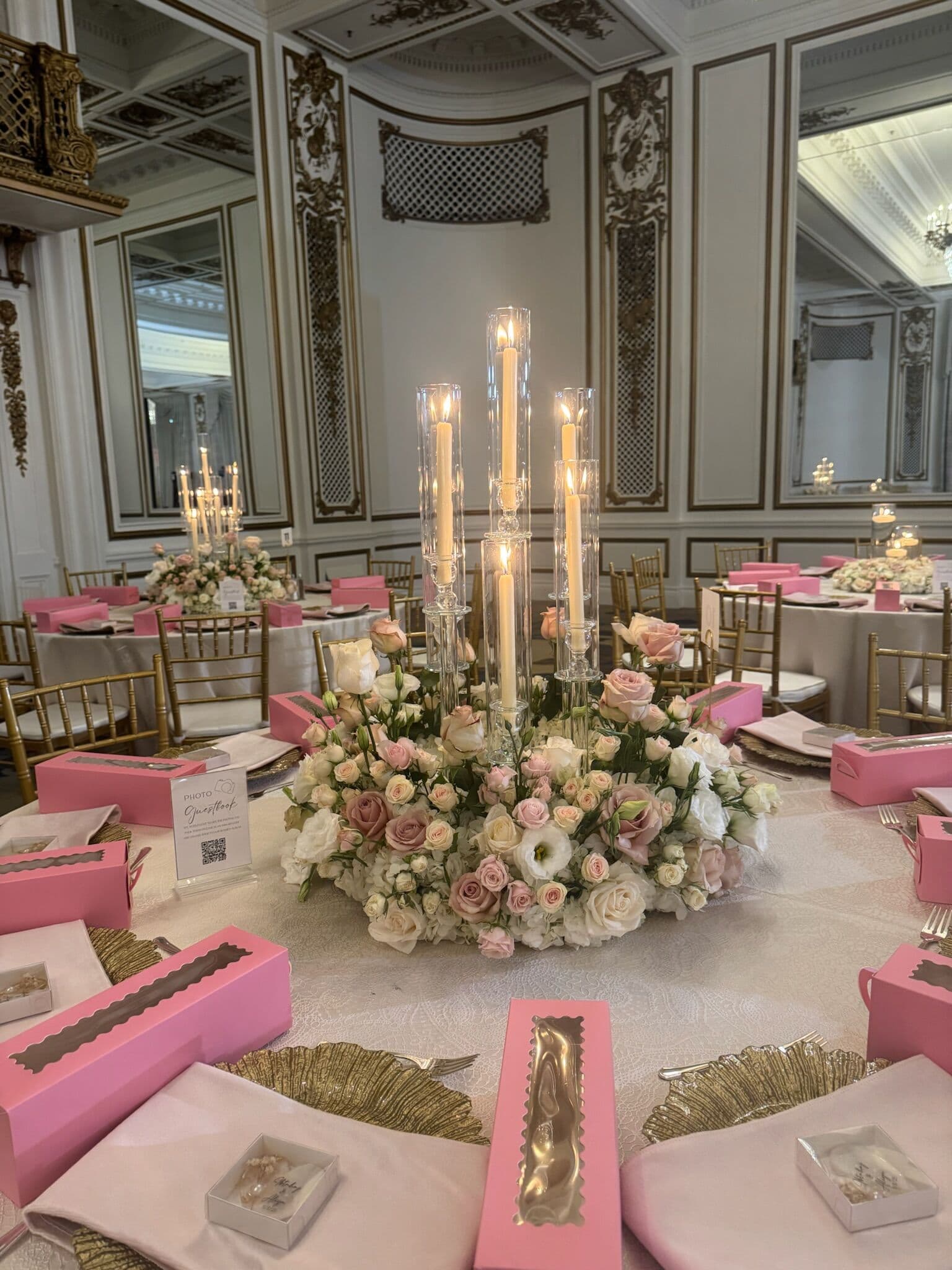 Ballroom centerpiece of blush and ivory roses beneath a candelabra of glass hurricane tapers, surrounded by pink napkins, gold leaf-shaped chargers and pink favor boxes in an ornate paneled Bay Area venue.