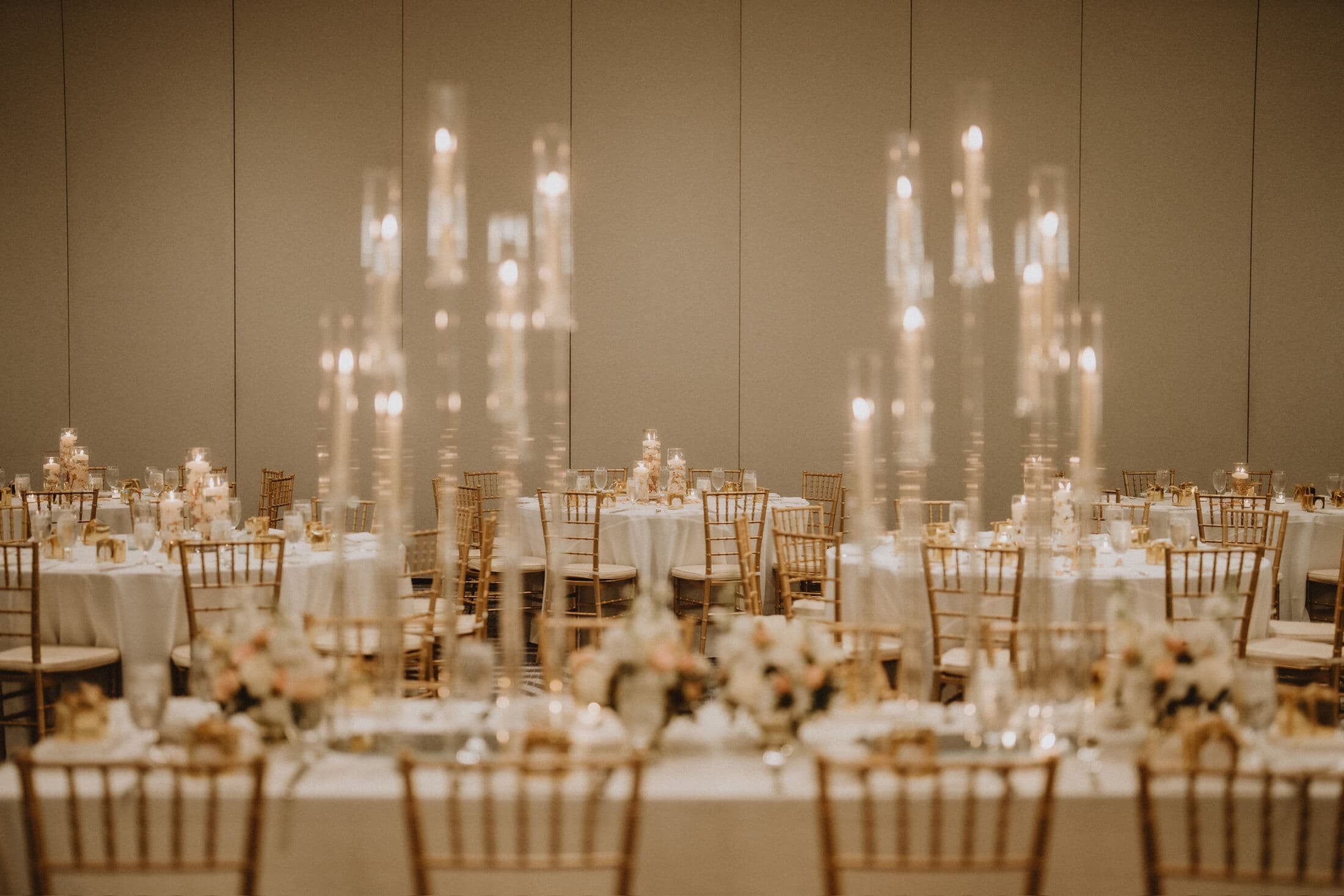 Ballroom reception in warm candlelight with white-linen round tables, gold Chiavari chairs, blush floral centerpieces and towering glass candelabra tapers glowing against a neutral wall.
