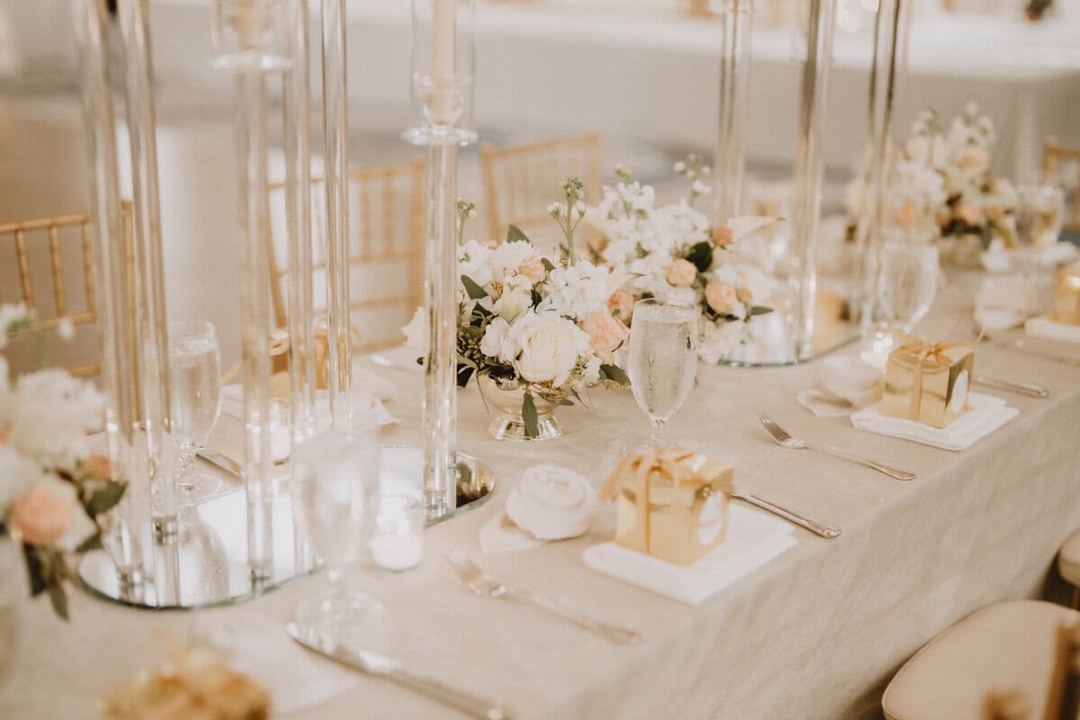 Head-table detail in soft neutral linen with tall glass candlesticks, a low centerpiece of white and peach roses and greenery, gold-ribboned favor boxes on folded napkins, water goblets and silver flatware.