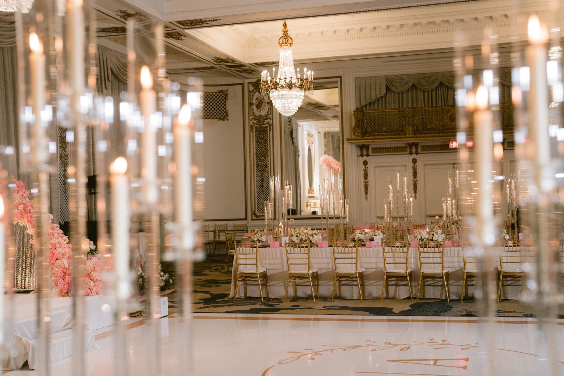 Grand ballroom with a crystal chandelier and gold-leaf paneling seen through a foreground of tall glass taper candles, a long head table lined with gold Chiavari chairs, pink accents and a blush floral entry arrangement.
