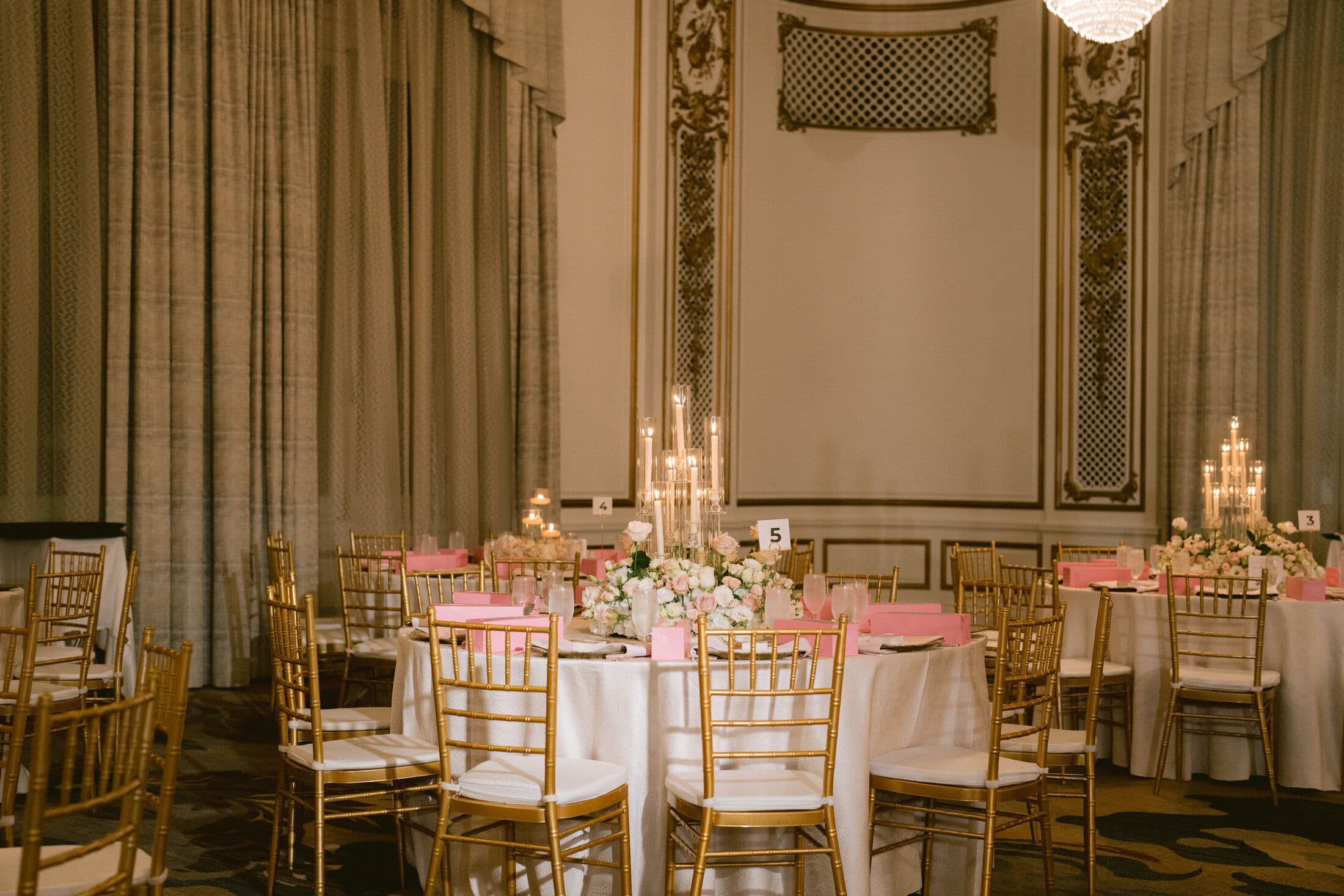 Elegant paneled ballroom with round tables in white linens, gold Chiavari chairs, pink napkins, candelabra tapers, blush-and-ivory floral centerpieces and a numbered table card beneath tall draped curtains.