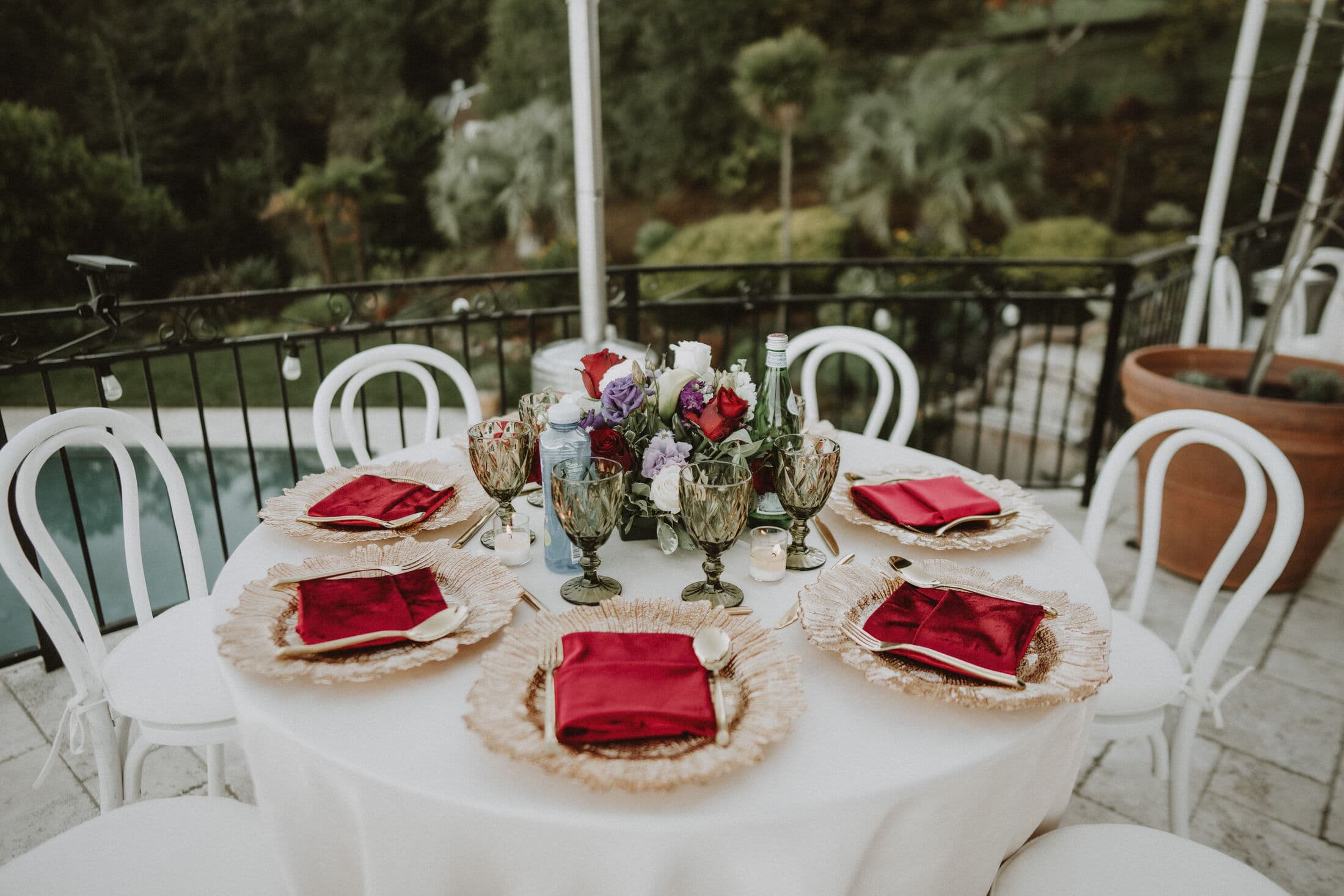 Round table on a balcony overlooking a pool and garden, set with gold-edged ruffled glass chargers, crimson satin napkins, smoke-gray goblets and a centerpiece of red and white roses with lisianthus among white bentwood chairs.