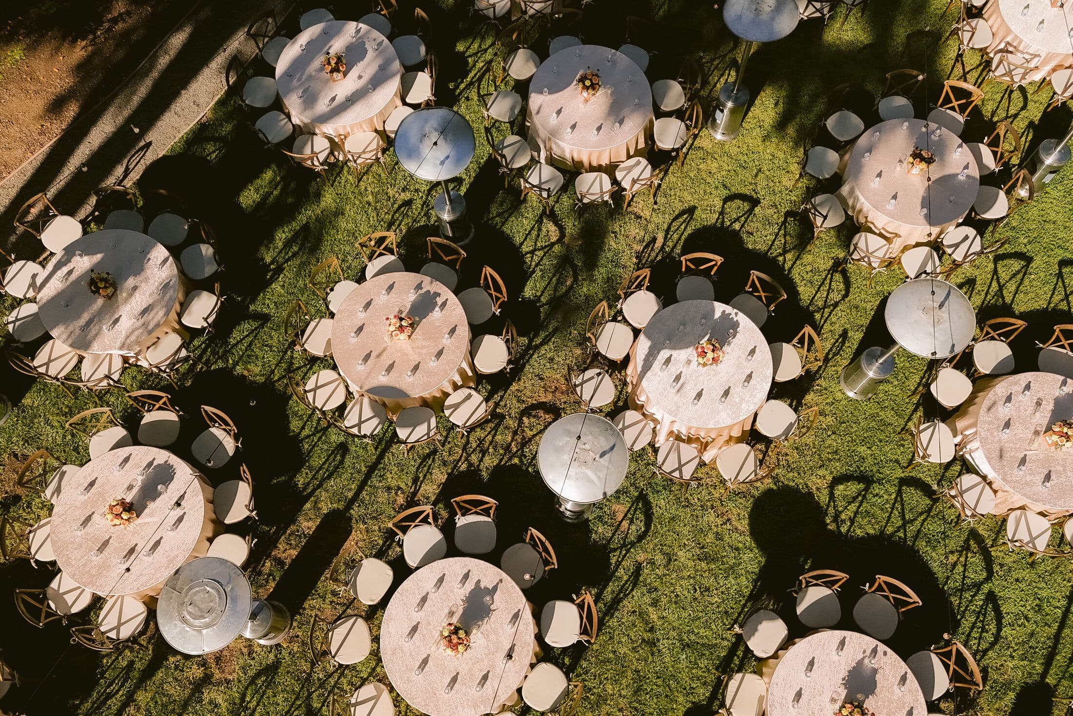 Aerial view of round guest tables in printed linens with cross-back chairs and small floral centerpieces, casting long shadows across a green lawn at an outdoor Bay Area event.