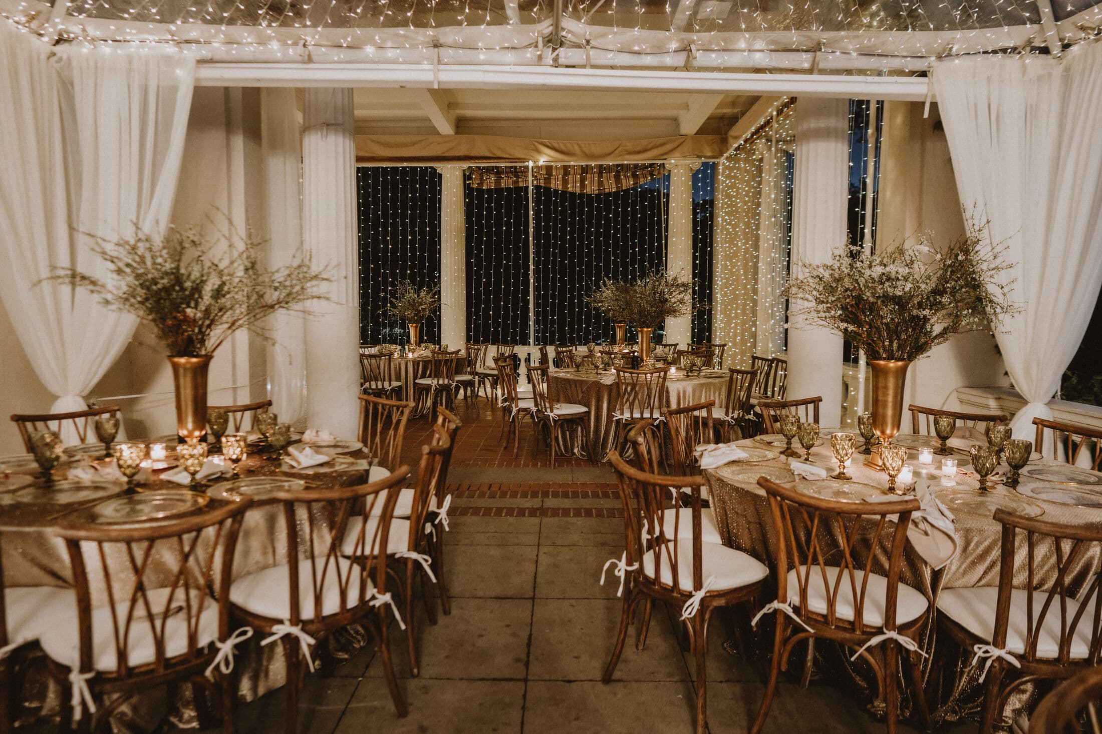 Draped patio reception lit by curtains of fairy lights, with round tables in shimmering linen, wooden cross-back chairs, votive candles and tall brass urns holding wispy dried-grass centerpieces.