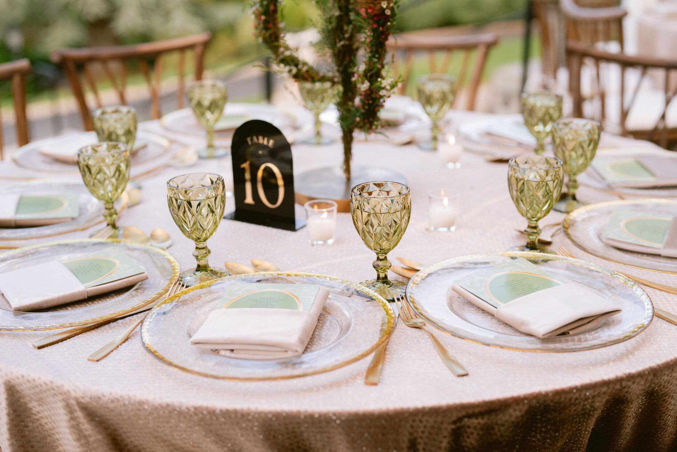 Round table in champagne sequined linen with gold-rimmed glass chargers, green sage napkins, gold flatware, faceted green goblets, a black arched 'Table 10' number and a tall greenery centerpiece at an outdoor Bay Area wedding.