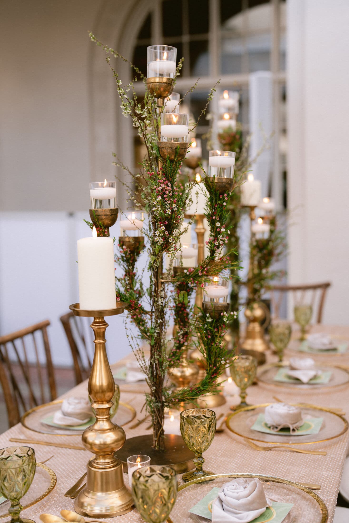 Long table centerpiece of brass pillar-candle stands and greenery branches with berry accents, flanked by sequined linen, glass chargers, green goblets and folded rosette napkins in a bright indoor Bay Area setting.