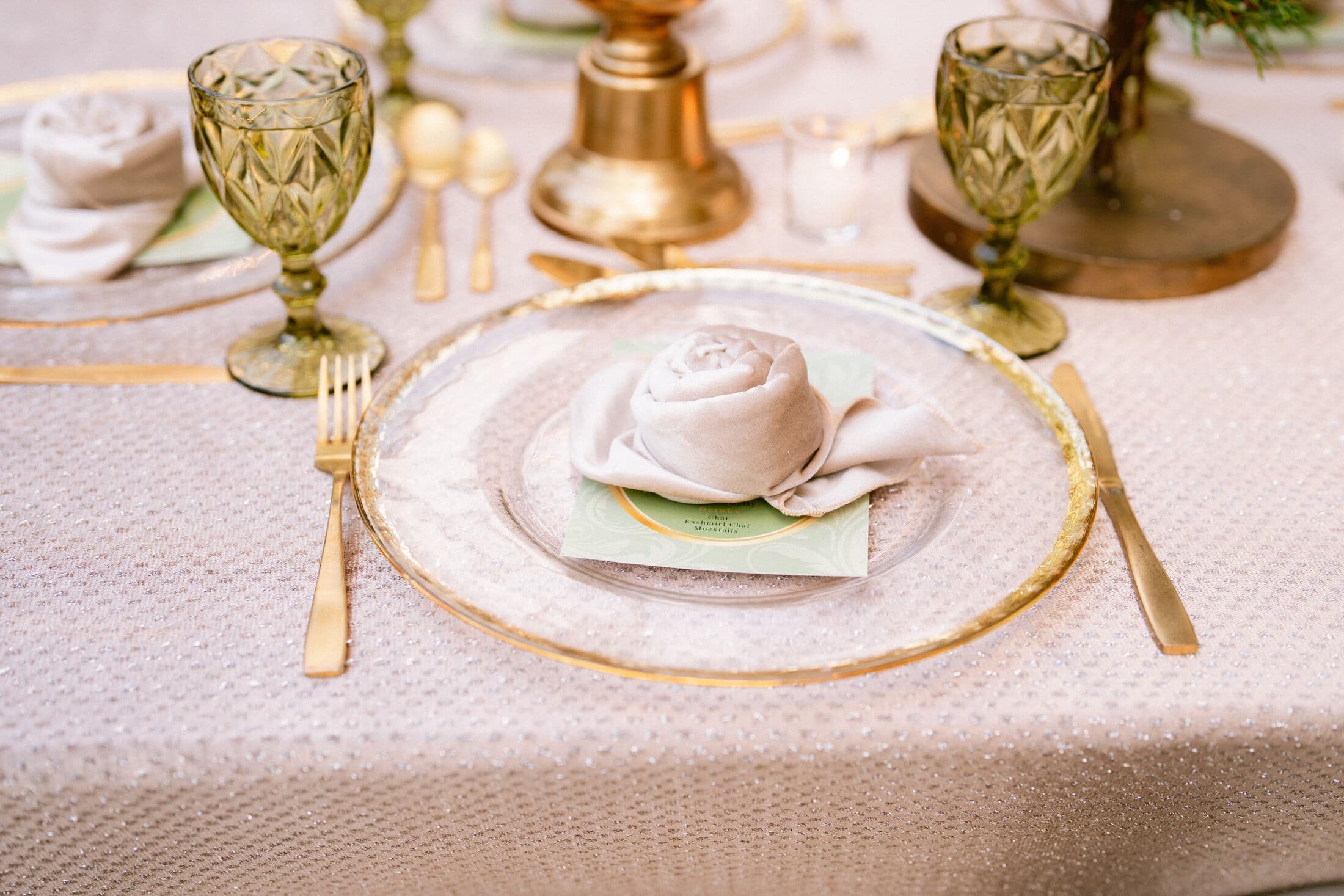 Place setting detail with a gold-rimmed clear glass charger, a rose-folded blush napkin on a sage green menu card, gold flatware and faceted green goblets atop shimmering champagne sequined linen.