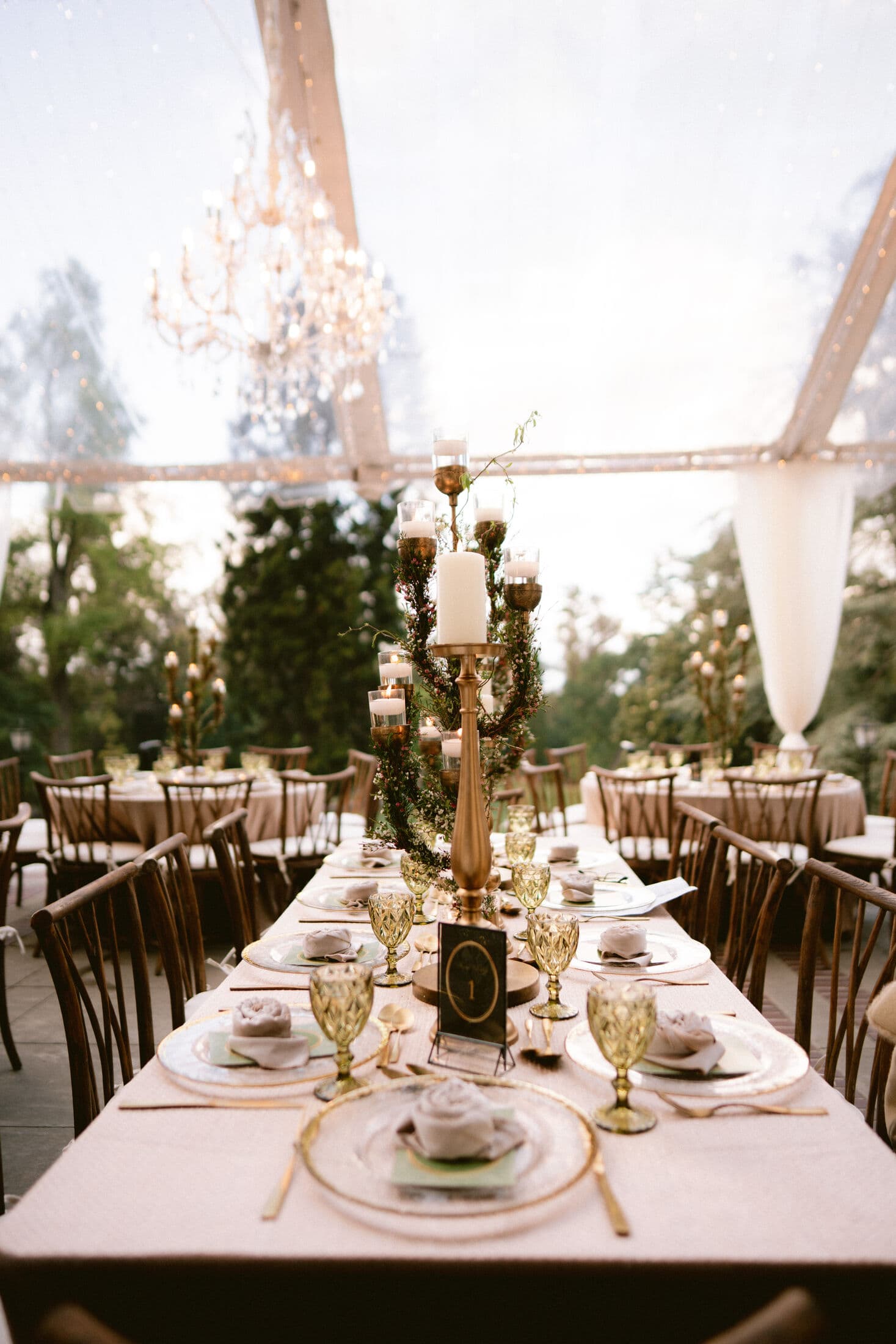 Clear-tented reception with crystal chandeliers overhead, a long blush-linen table set with gold-rimmed chargers, rose-folded napkins, green goblets, a black 'Table 1' number and a brass candelabra with greenery, ringed by wooden cross-back chairs.