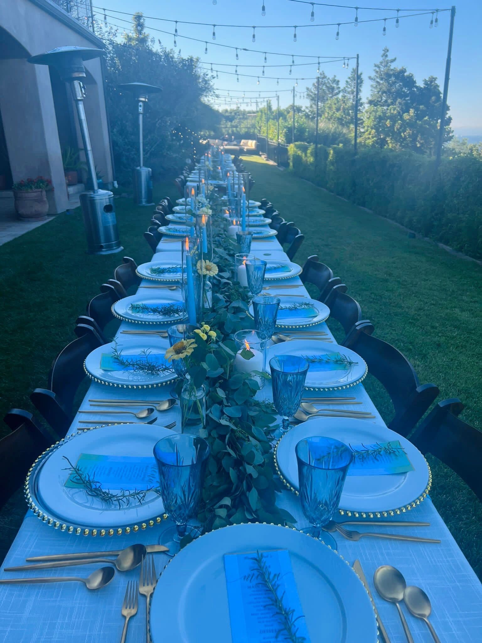 Long banquet table at golden hour in the Bay Area hills with gold-beaded chargers, white plates, blue goblets, gold flatware, sunflower and eucalyptus accents and a eucalyptus runner, lined with dark chairs by Memorable Moments by Ladle n Wok.
