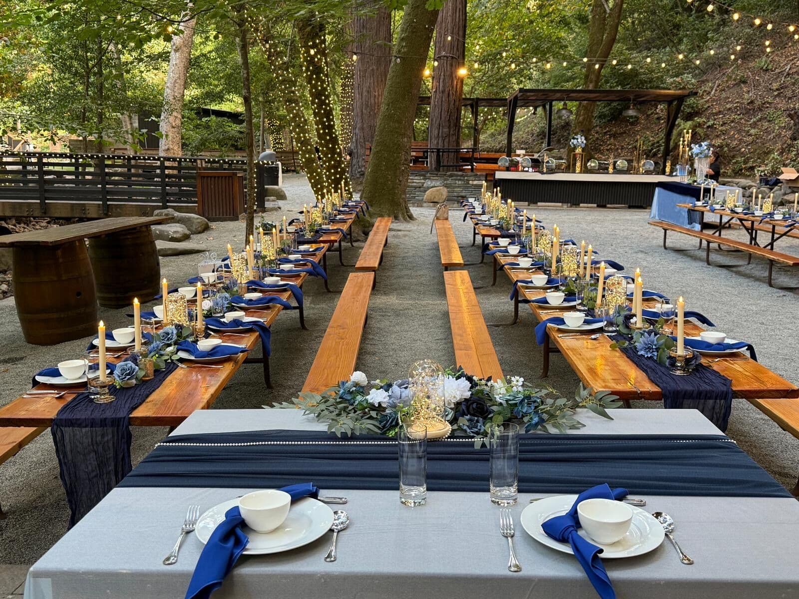 Rustic outdoor table layout with wooden picnic-style tables, gray runners, navy napkins folded in white bowls, gold taper candles and blue-and-white floral centerpieces, set among redwoods under string lights.