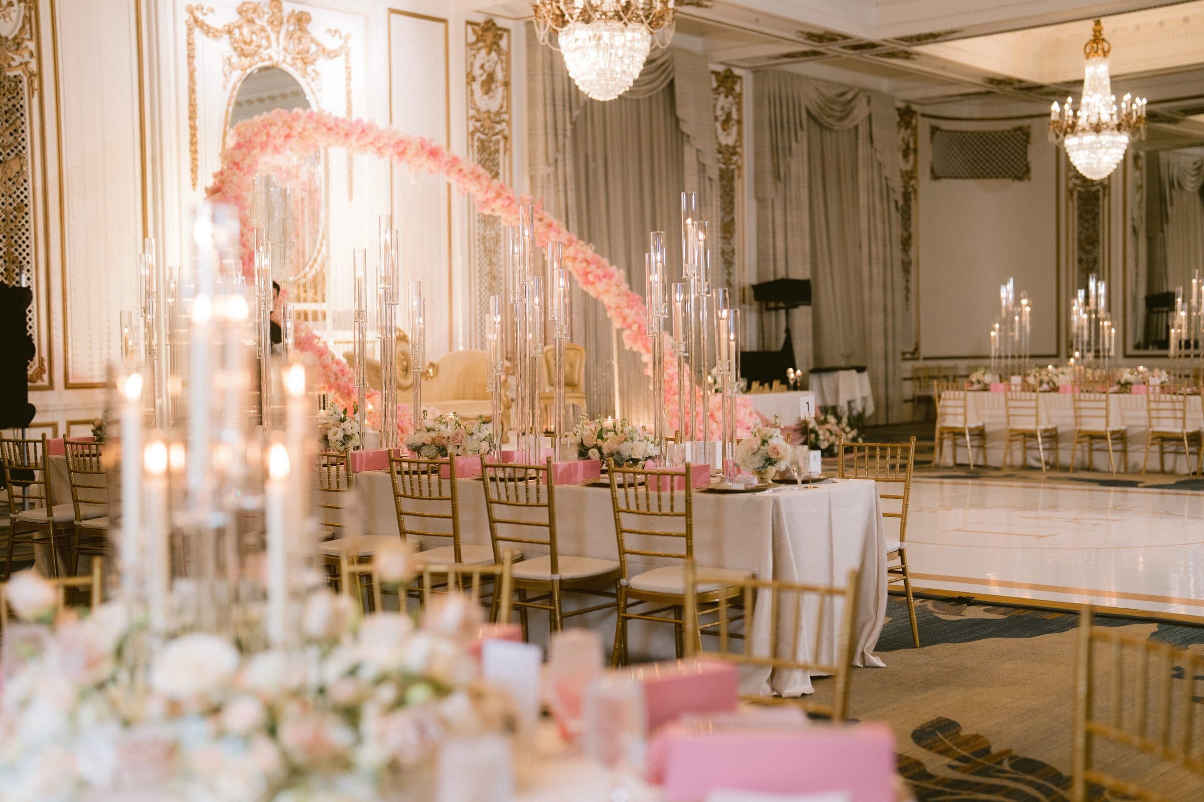 Grand ballroom set for a walima — a sweeping pink-and-blush floral arch curving over a gilded settee, seen down a long head table of tall crystal candelabras, blush rose centerpieces and gold Chiavari chairs, with crystal chandeliers above and the dance floor beyond.