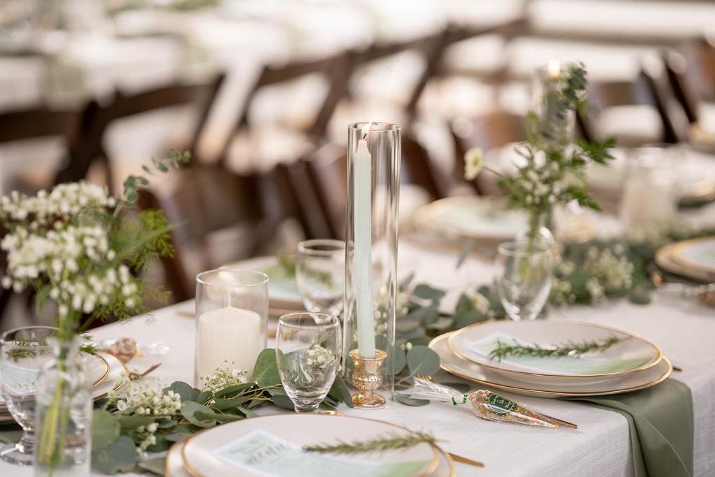 Close detail of a sage-and-ivory place setting — a mint taper burning in a tall glass hurricane on a gold holder, a pillar candle, cut-glass goblets, eucalyptus and baby's breath trailing along the linen, and a gold-rimmed plate set with a rosemary sprig on a printed menu card.